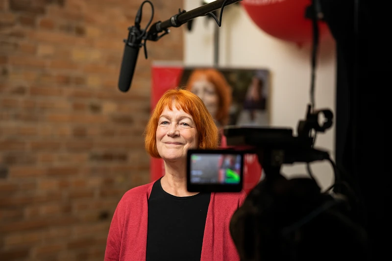 Imagevideo Produktion 1 Professionelles Imagefilm Interview mit lächelnder Frau (rotes Haar, roter Cardigan) vor Backsteinwand. Mikrofon und Kamera-Display im Vordergrund.